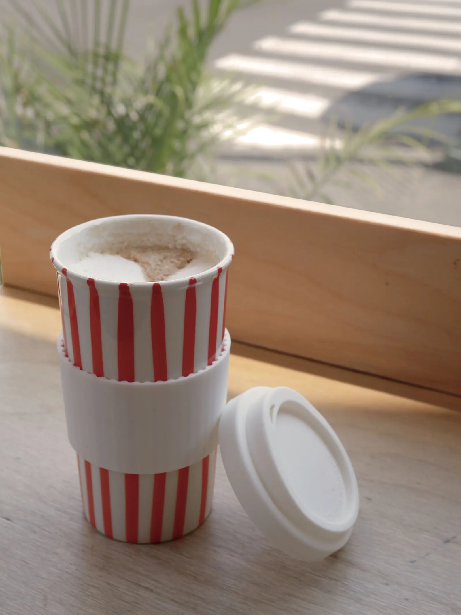 Red striped Millie cup with latte, silicone sleeve and lid on a windowsill